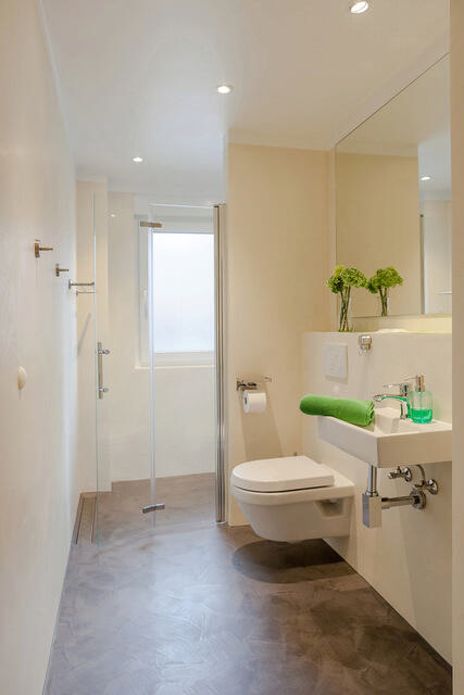 Ein modernes Einfamilienhaus in einer wohlgeordneten Umgebung. Das Haus hat eine klassische weiße Fassade mit einem roten Ziegeldach. Das Giebelfenster und die zwei kleineren Dachfenster lassen viel natürliches Licht ins Innere. Der gepflegte Vorgarten ist mit verschiedenen Pflanzen und Sträuchern bepflanzt, die einen Kontrast zur Einfahrt aus grauen Pflastersteinen bilden. Es gibt auch eine Garage auf der rechten Seite, die gut in das Gesamtbild des Hauses integriert ist. Der Eingangsbereich ist durch einen kleinen Gehweg, flankiert von dekorativen Pflanzen, zugänglich. Insgesamt strahlt das Haus eine gemütliche und einladende Atmosphäre aus, ideal für eine Familie.